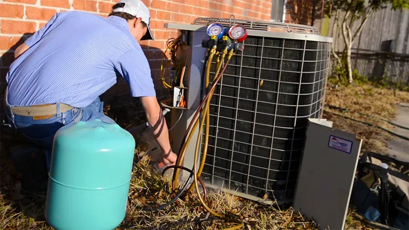 A technician kneeling at an outdoor cooling condenser with a red-and-blue gauge manifold connected to the service ports