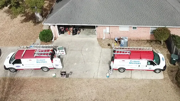 Two branded Climate Masters vans in a Summerdale driveway, with a technician working at the outdoor unit at the side of a brick home