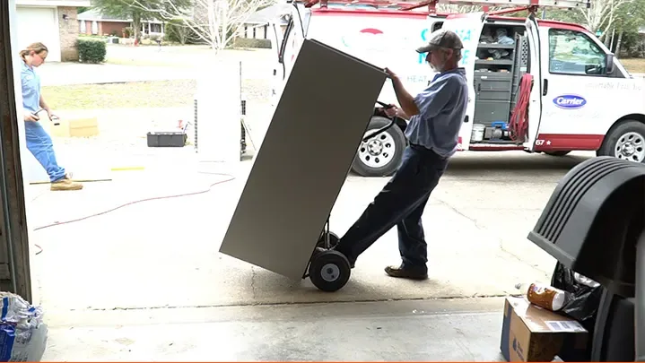 A technician wheeling a tall air handler cabinet on a hand truck up a driveway, with a Climate Masters service van parked behind
