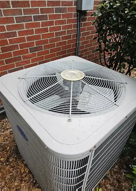 A white Carrier outdoor condensing unit standing against a red brick wall, seen from above.