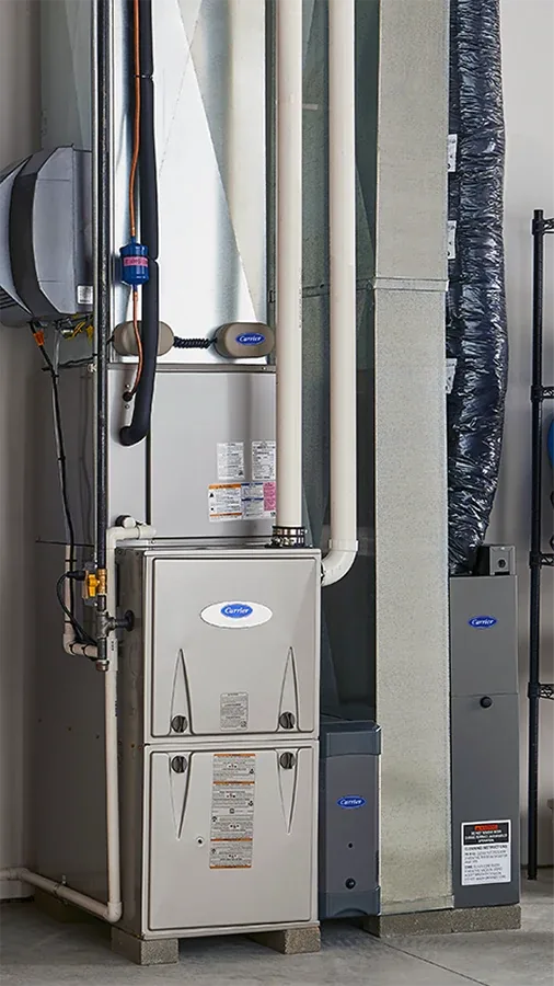 A Carrier gas furnace standing in a narrow indoor utility space beside its sheet-metal supply ductwork and an insulated flexible duct run