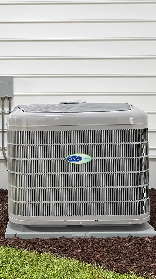 A Carrier heat pump unit standing on its pad outside a home with white siding.