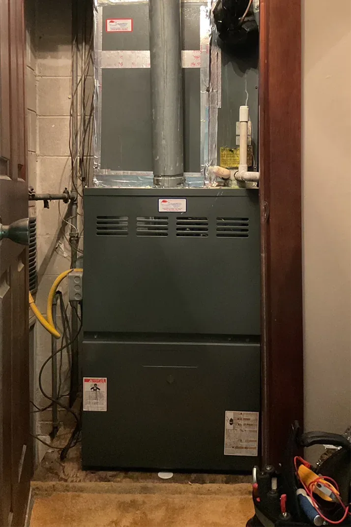 An older dark furnace standing in a basement doorway with a technician's tool bag beside it