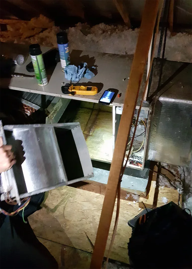 A technician working on ductwork in an attic, with a meter and sealant on a board beside the opening
